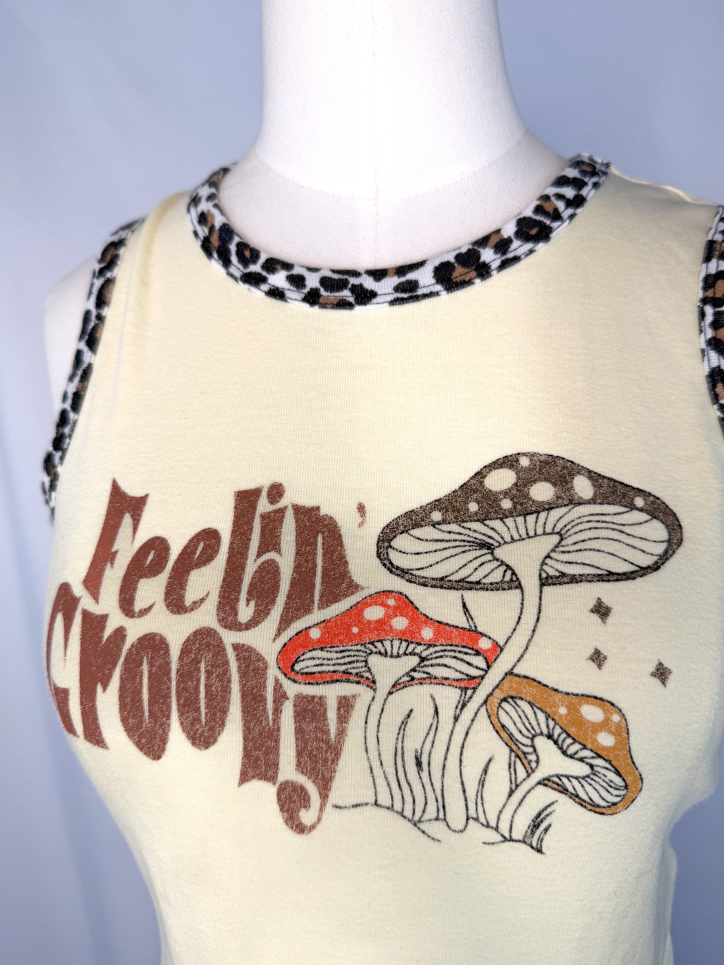 Up-close shot of Urban Outfitters Feeling Groovy Crop Top in Yellow. Focusing on the "Feelin' Groovy" letters next to three mushrooms and the cheetah print detail on the neckline and arm cutouts.