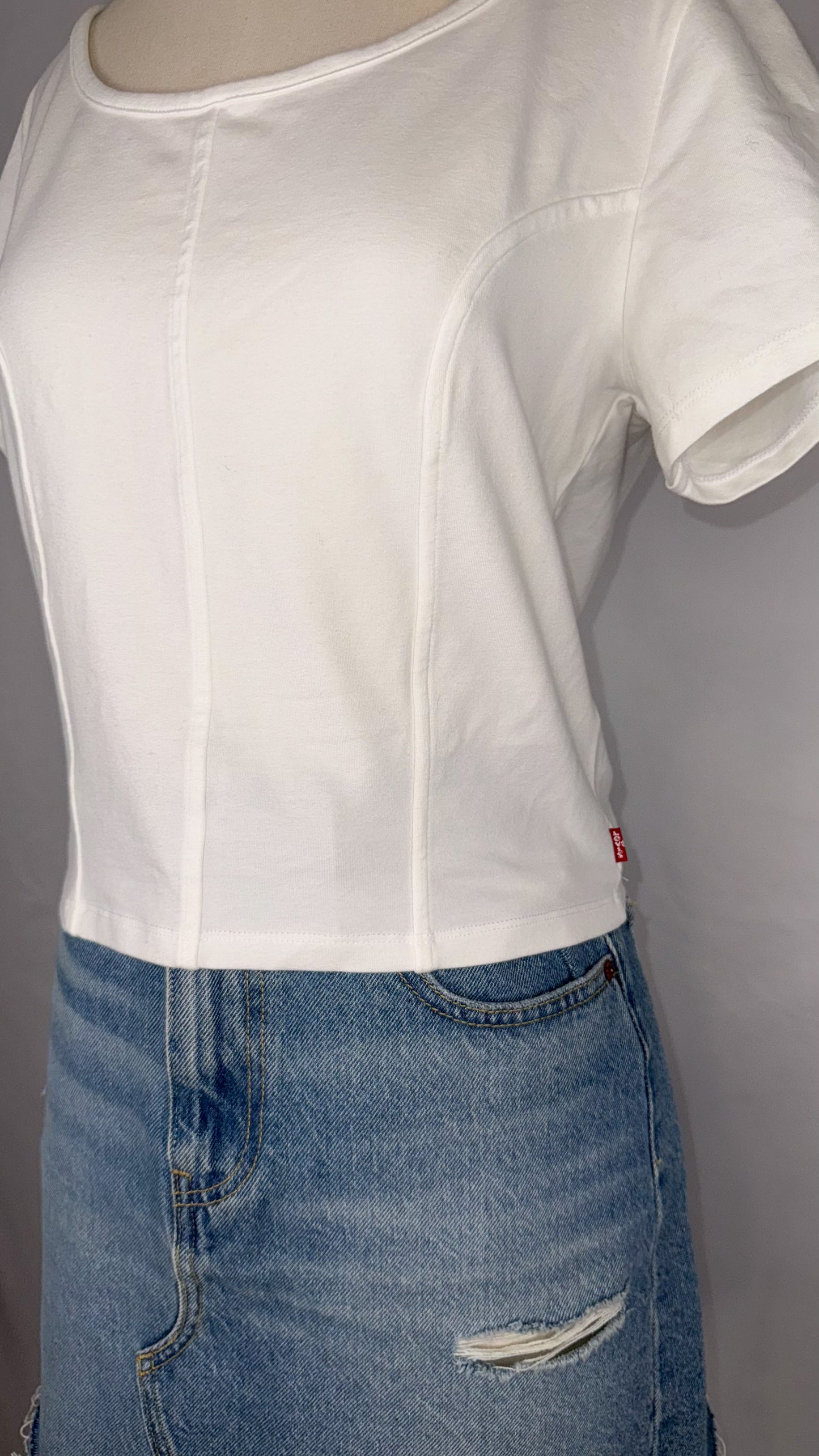 Up-close side view of Levi's Corset T-Shirt in White featuring the small red Levi's tag on the right side of the shirt, as well as the corset style stitching. Mannequin is wearing the top paired with the distressed light blue denim mini skirt.