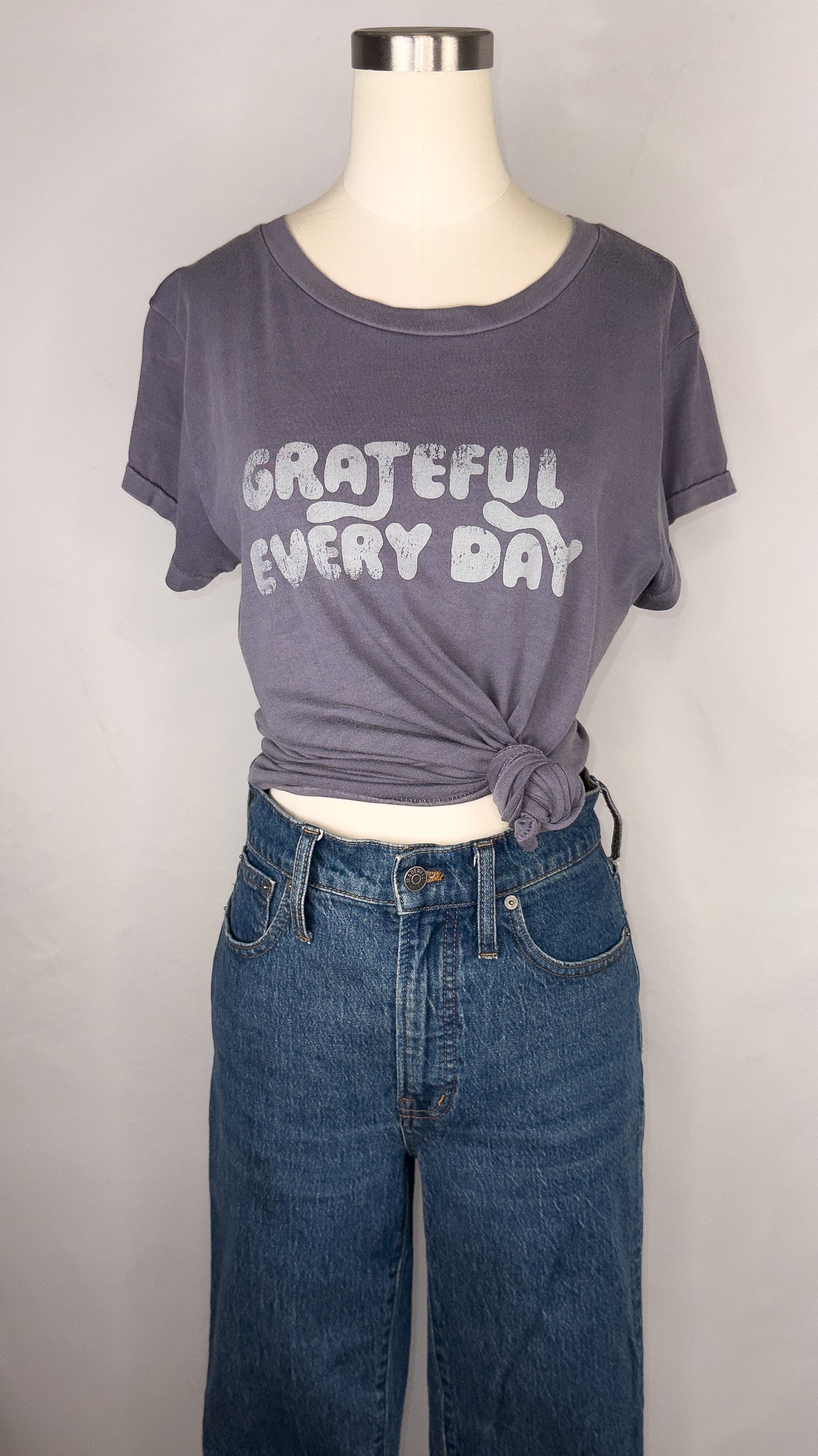 Front facing view of Rivet & Thread Grateful Everyday Graphic Ex-Boyfriend Tee in Sunfaded Indigo. The t-shirt is styled on the mannequin with a knot to the side, and a pair of dark blue jeans. Across the front of the shirt reads "Grateful Every Day" in faded-ish letters.