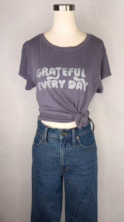 Front facing view of Rivet & Thread Grateful Everyday Graphic Ex-Boyfriend Tee in Sunfaded Indigo. The t-shirt is styled on the mannequin with a knot to the side, and a pair of dark blue jeans. Across the front of the shirt reads "Grateful Every Day" in faded-ish letters.