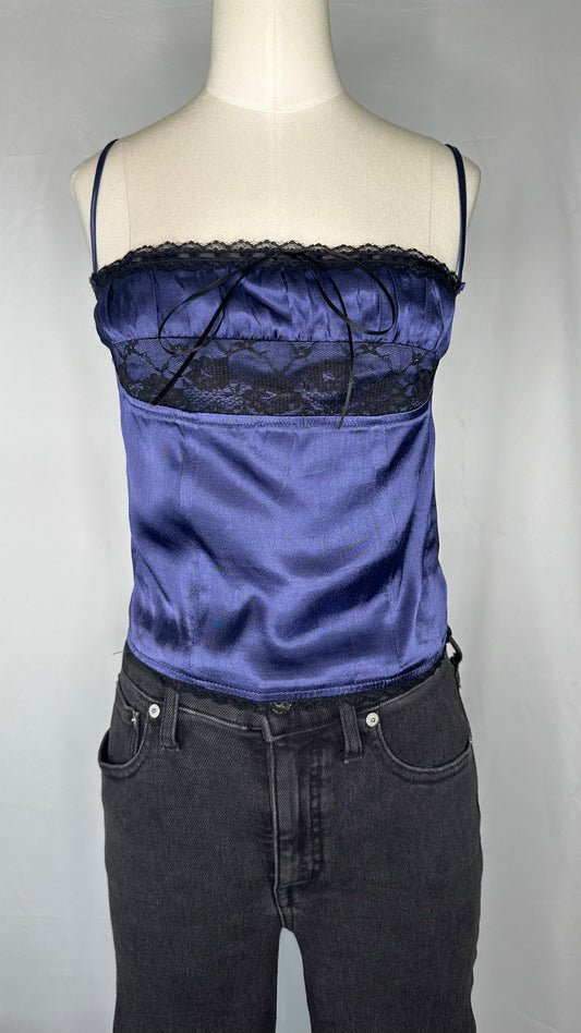 Front view of Sky to Moon Satin Lace Cami Top in Navy, featuring the lace detail along the top & bottom of the cami, a bow in the middle, and spaghetti straps. Mannequin is shown wearing the cami paired with black denim jeans.  