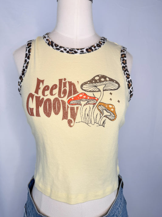 Front shot of Urban Outfitters Feeling Groovy Crop Top in Yellow, featuring a cheetah prink neckline and outline on arm cutouts. "Feelin' Groovy" in funky letters next to three mushrooms of different colors.